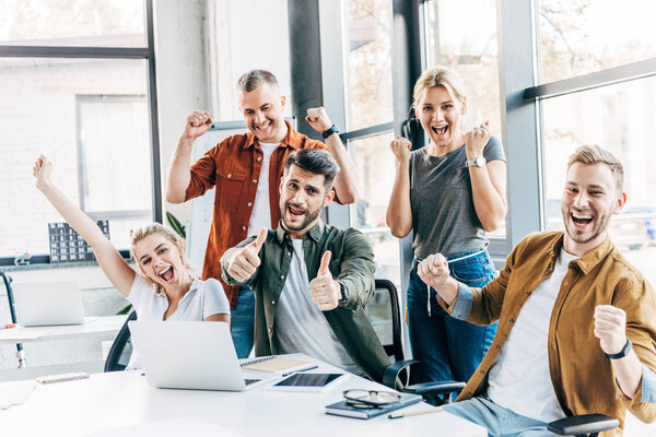 группа счастливых успешных предпринимателей, празднующих победу и показывающих большие пальцы в офисе и смотрящих в камеру
