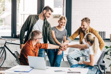 group of smiling young entrepreneurs making team gesture while working on startup together at office clipart