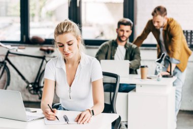 dizüstü bilgisayar ve Not defteri ile ofiste meslektaşları ile arka planda çalışan güzel bir genç kadın