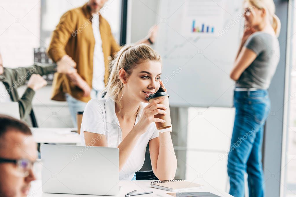 smiling young woman talking by phone and drinking coffee at office with colleagues on background