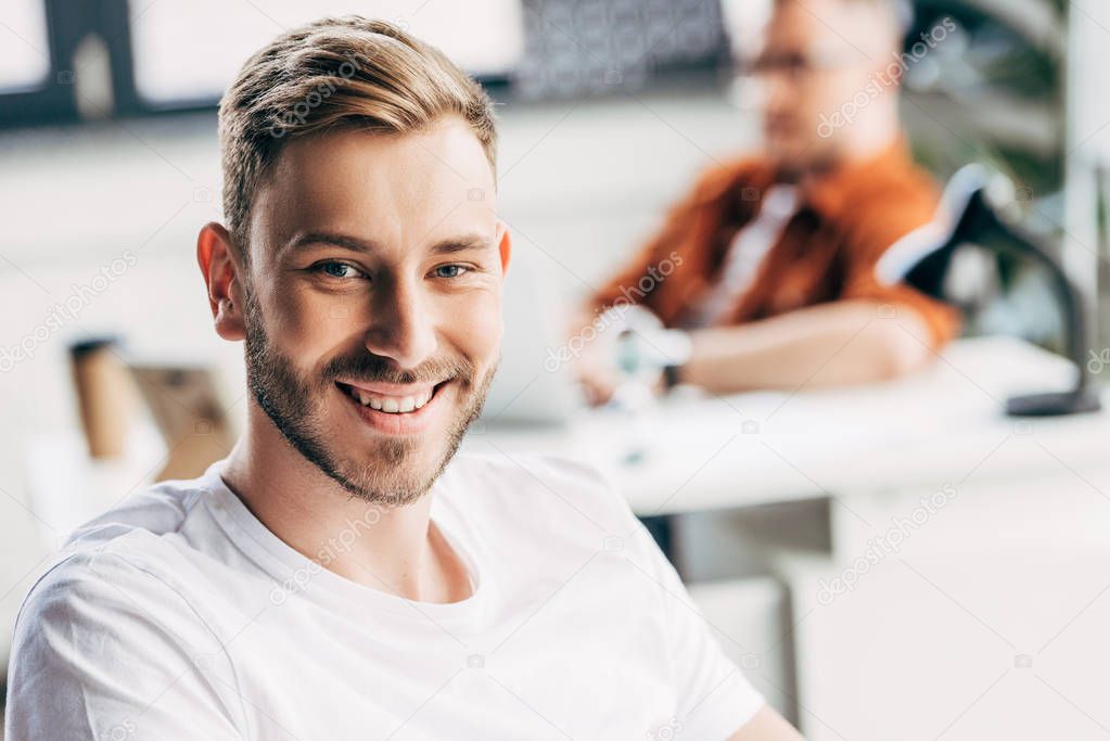 handsome young businessman looking at camera with colleague sitting blurred on background at office