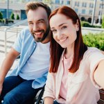 Camera point of view of happy boyfriend in wheelchair and beautiful girlfriend looking at camera in city