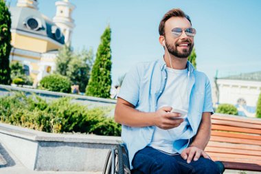 uzağa arıyorsunuz gülümseyen yakışıklı adam akıllı telefon tutarak ve müzik bir sokakta, tekerlekli sandalyede