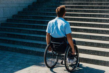 back view of man using wheelchair on street and looking at stairs without ramp clipart