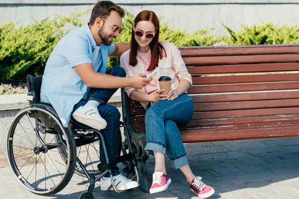 英俊的男朋友在轮椅和女朋友看在街上的智能手机 — 图库照片