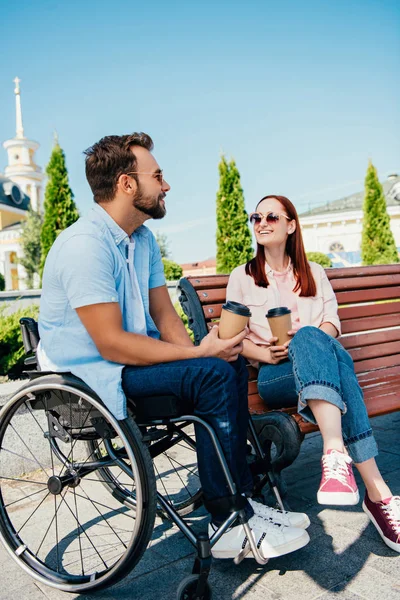 cheerful boyfriend in wheelchair and girlfriend with disposable coffee cups on street