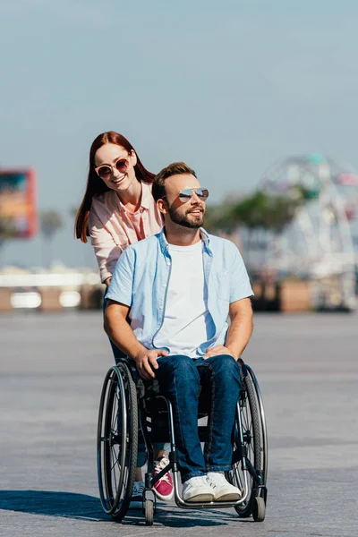 迷人的微笑的女朋友推英俊的男朋友在轮椅上街头 — 图库照片