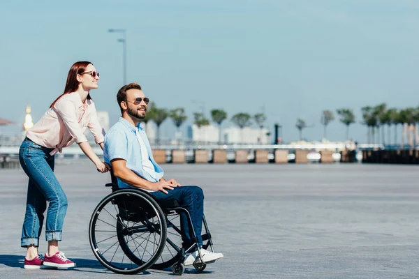 侧面视图的微笑迷人的女朋友推英俊的男朋友在轮椅上街头 — 图库照片