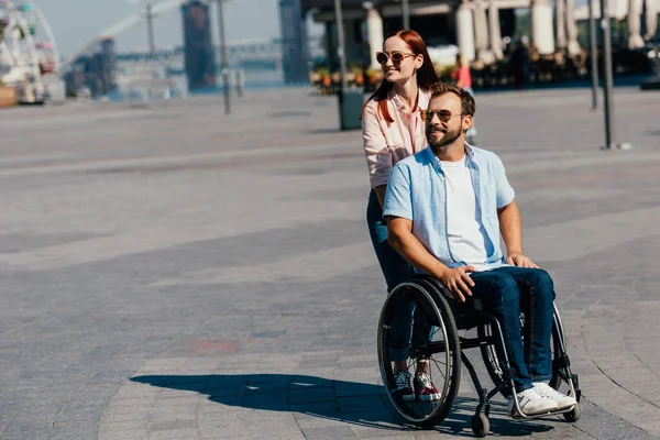 有吸引力的女朋友在太阳眼镜推残疾人英俊的男朋友在轮椅上街头 — 图库照片