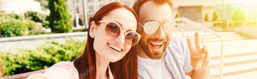 panoramic view of happy boyfriend and girlfriend in sunglasses looking at camera in city, man showing peace sign