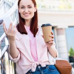 Çekici Dişileştirilmiş kadın smartphone tek kullanımlık kahve fincanı ile tutarak ve sokakta kameraya bakarak pembe gömlekli