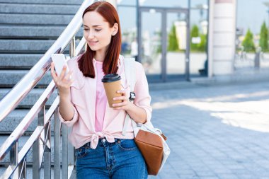 attractive redhair woman in pink shirt using smartphone and holding disposable coffee cup on street clipart