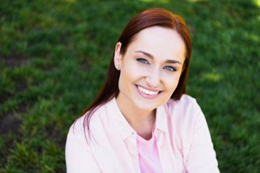 portrait of attractive redhair woman in pink shirt looking at camera in park clipart