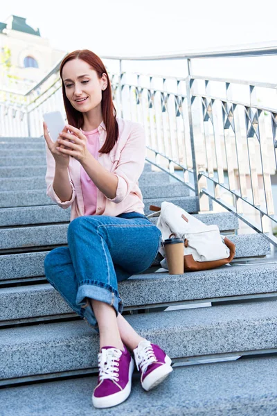 低角度视图的微笑迷人的鲜红妇女在粉红色衬衫坐在楼梯上 使用智能手机 — 免费的图库照片