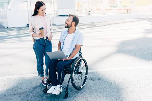 handsome boyfriend in wheelchair using laptop and girlfriend standing with coffee in paper cup on street, looking at each other