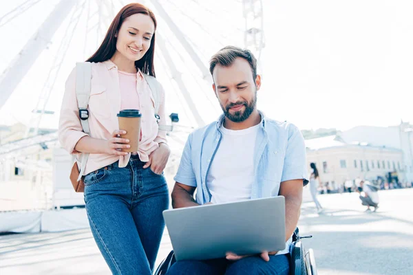 handsome boyfriend in wheelchair using laptop and girlfriend standing with coffee in paper cup on street