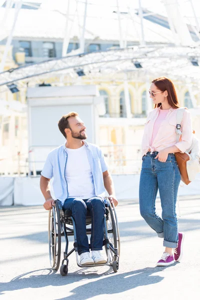 微笑英俊的男朋友在轮椅和女朋友看着对方在街上 — 免费的图库照片