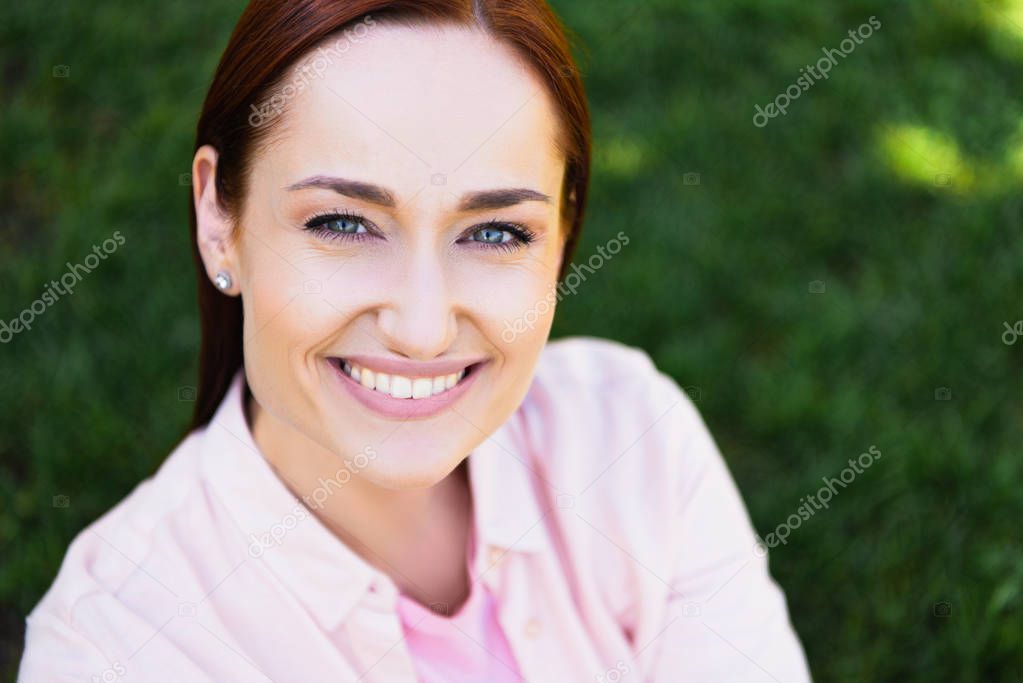 head and shoulders of attractive happy redhair woman in pink shirt looking at camera in park