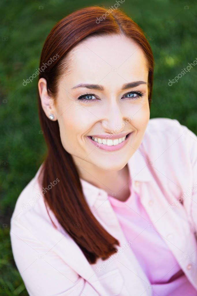 high angle view of attractive smiling redhair woman in pink shirt looking at camera in park