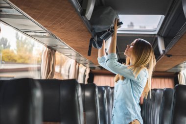 side view of young woman putting backpack on shelf in travel bus clipart