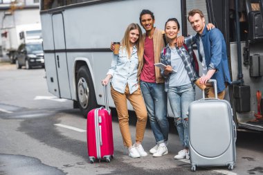 young happy multicultural friends with wheeled bags posing near travel bus at street  clipart