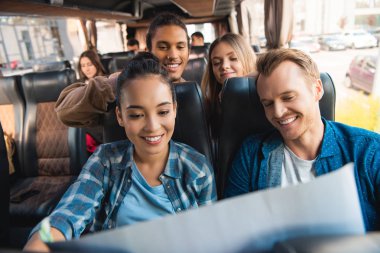 cheerful multiethnic tourists looking at map during trip on travel bus clipart