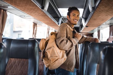 smiling multiracial male tourist with backpack looking at camera in travel bus clipart