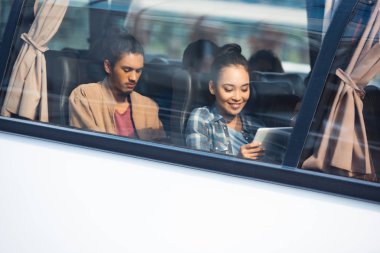 gülümseyen Asyalı kadın karışık yarış erkek Seyahat Otobüs gezisi sırasında arkasında oturan arkadaşı ise dijital tablet kullanma