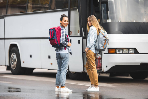 young multiethnic women with backpacks posing near travel bus at urban street