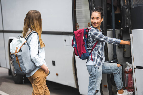 multicultural female travelers with backpacks walking into travel bus at street