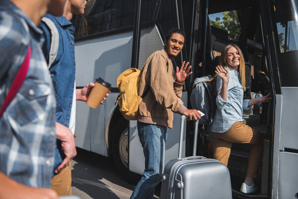 happy multiethnic tourists with backpacks and wheeled bags waving by hands to friends while walking into bus at street