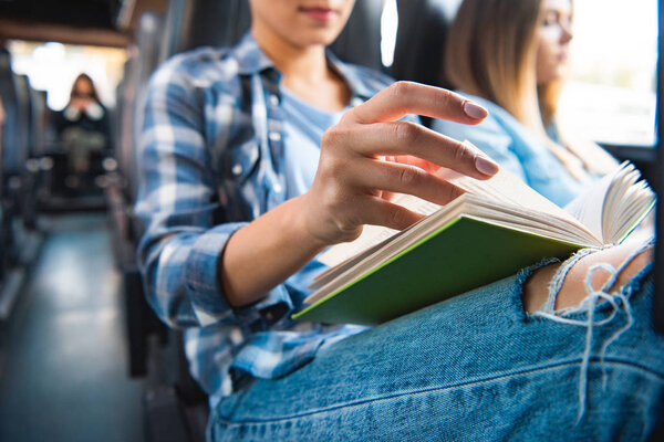 cropped image of woman reading book while her female friends sitting near during trip on travel bus