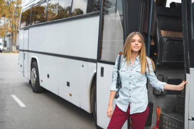 young woman with rucksack standing near travel bus at street clipart