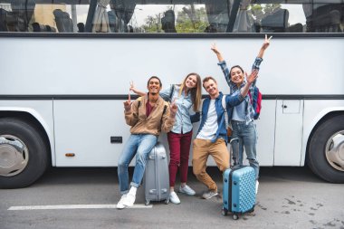 cheerful multiethnic tourists with travel bags doing peace signs near bus at street clipart