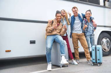 smiling multiethnic friends with wheeled bags doing thumbs up and peace gestures near travel bus clipart