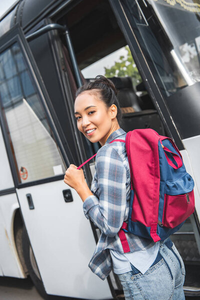 selective focus of asian female tourist with backpack looking at camera near travel bus at city street