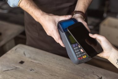 cropped shot of waiter holding payment terminal while customer doing contactless purchase with smartphone in cafe clipart