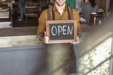 cropped shot of smiling young waiter holding chalkboard with open inscription in cafe clipart
