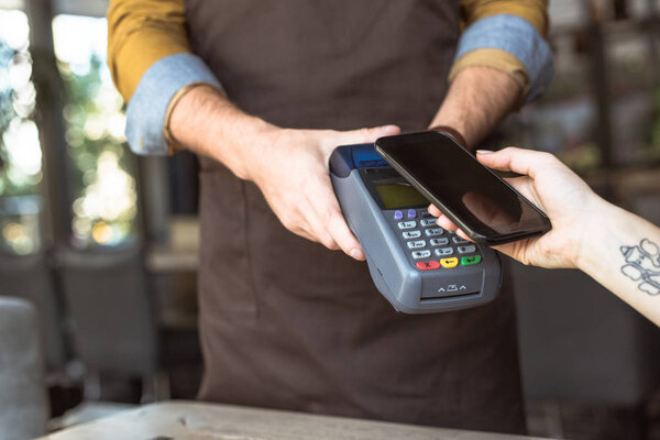cropped shot of waiter holding payment terminal while customer doing contactless purchase with smartphone in cafe