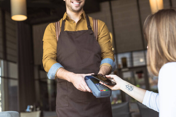 cropped shot of smiling waiter holding payment terminal while customer doing contactless purchase with smartphone in cafe