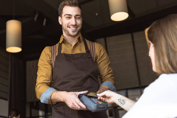 smiling young waiter holding payment terminal while customer doing contactless purchase with credit card in cafe