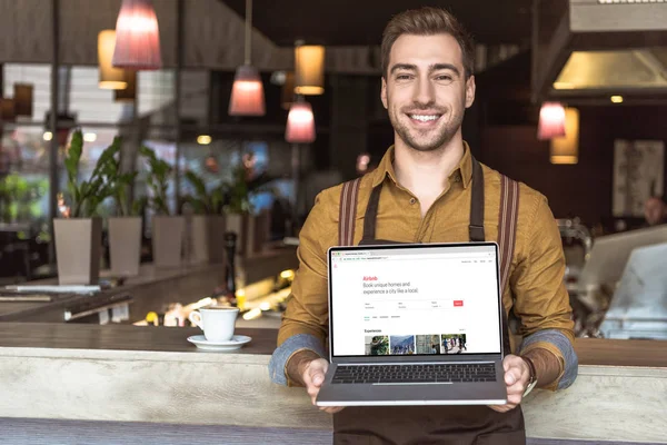 Przystojny Młody Kelner Trzymając Laptop Strony Airbnb Ekranie Kawiarni — Zdjęcie stockowe