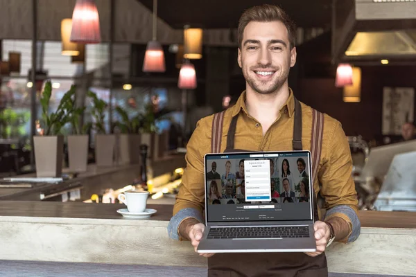 英俊的年轻侍者手持笔记本电脑与领英网站在屏幕上的咖啡厅 — 图库照片