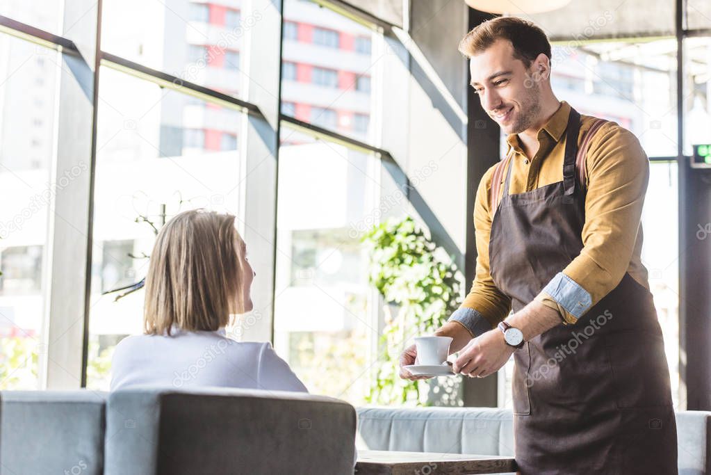 attractive young waiter serving cup of coffee for female client at cafe