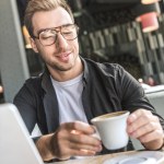 Primer plano de atractivo joven freelancer con portátil sosteniendo taza de café en la cafetería