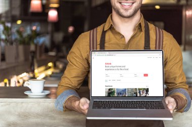 airbnb Web sitesi ile dizüstü bilgisayar ekranında kafede tutan gülümseyen genç garson kırpılmış atış