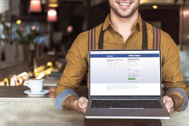 Web sitesi facebook ile dizüstü bilgisayar ekranında kafede tutan gülümseyen genç garson kırpılmış atış