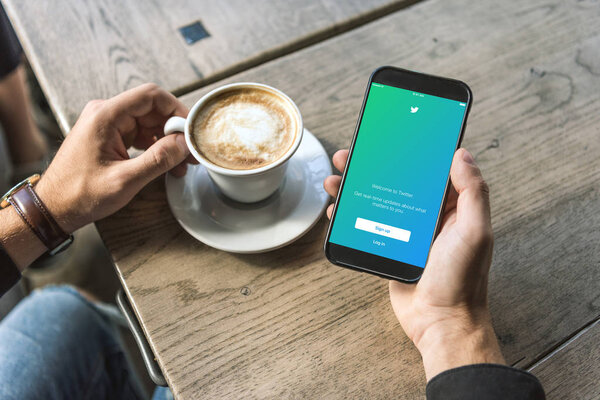 cropped shot of man with cup of cappuccino using smartphone with twitter app on screen