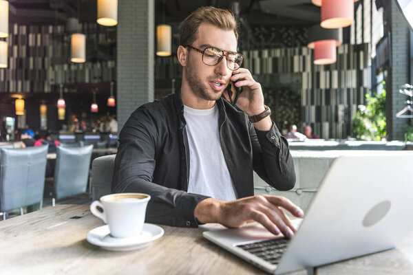 handsome young freelancer with cup of coffee working with laptop in cafe and talking by phone