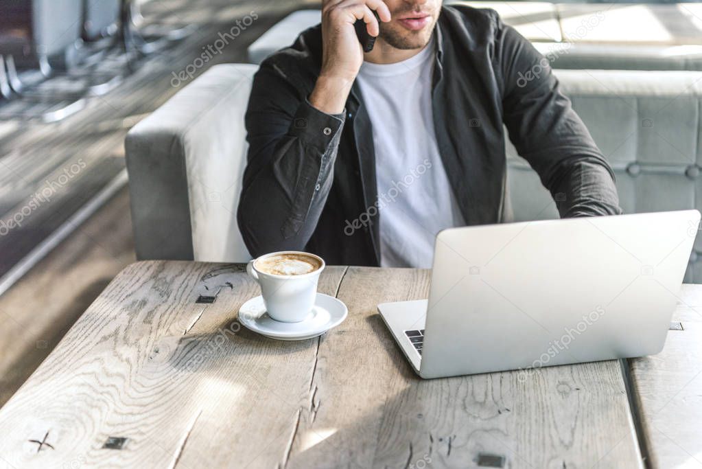 cropped shot of young freelancer working with laptop and talking by phone in restaurant
